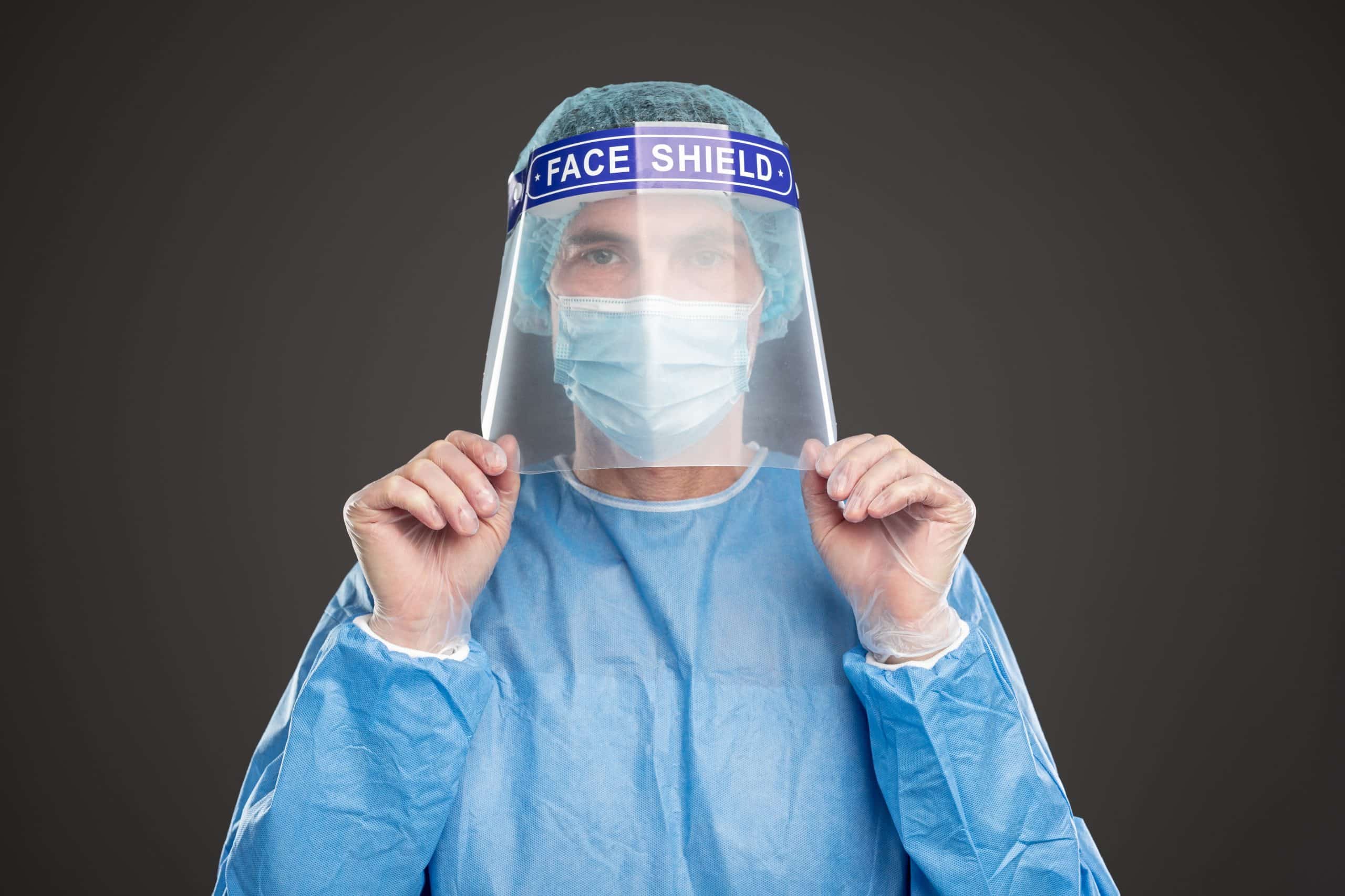 Male medic wearing protective suit and plastic face shield during COVID 19 outbreak while standing on gray background and looking at camera